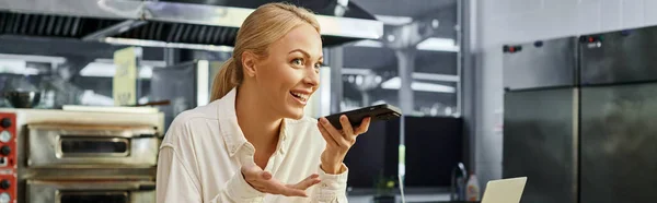 Femme élégante positive envoyant un message vocal sur smartphone dans un café confortable moderne, bannière horizontale — Photo de stock