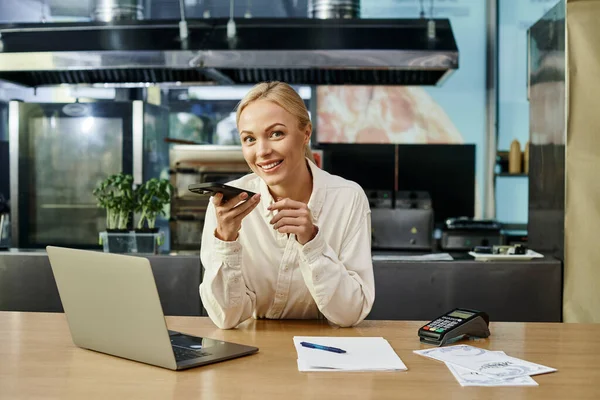Blonde woman sending voice message on smartphone and smiling at camera near laptop in cafe — Stock Photo