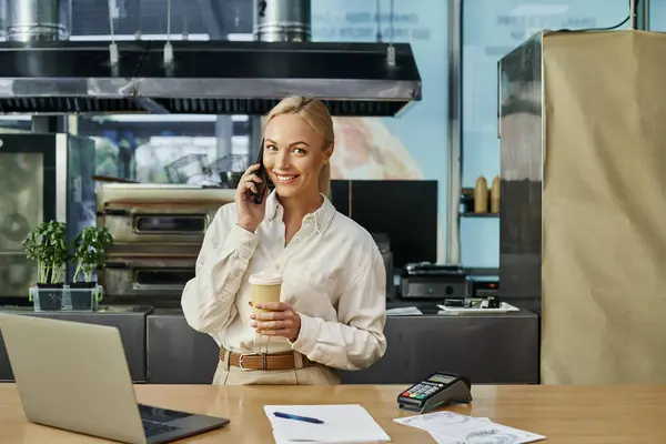 Felice amministratore caffè con caffè per andare a parlare su smartphone vicino laptop e terminale di pagamento — Foto stock