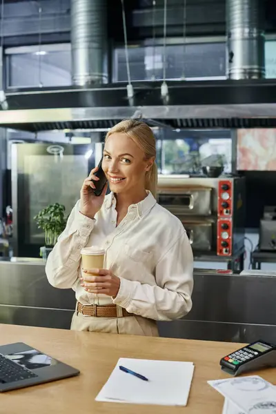 Gerente de café rubia feliz con café para ir a hablar en el teléfono inteligente cerca de la computadora portátil y terminal de pago - foto de stock