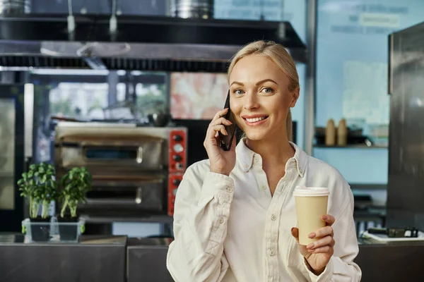 Gerente de café loira alegre com café para ir falar no smartphone e sorrindo para a câmera — Fotografia de Stock