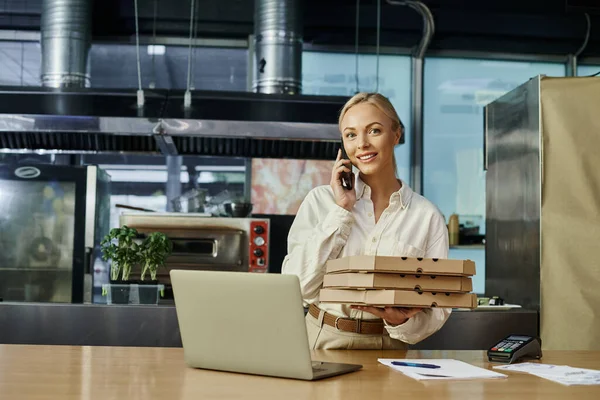Happy pretty manager with pizza boxes talking on smartphone near payment terminal and laptop in cafe — Stock Photo