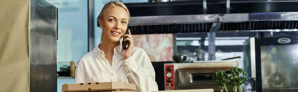 Sonriente rubia gerente de café con cajas de pizza hablando en el teléfono móvil y mirando a la cámara, pancarta - foto de stock