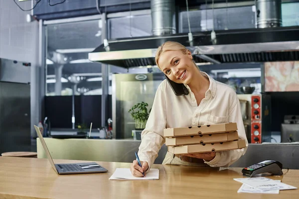 Administrador sonriente con cajas de pizza hablando en el teléfono inteligente y el orden de escritura en el mostrador en la cafetería - foto de stock