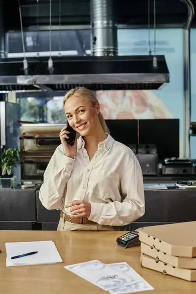 Gerente mujer sonriente hablando en el teléfono inteligente cerca de cajas de pizza y terminal de pago en el mostrador de café - foto de stock