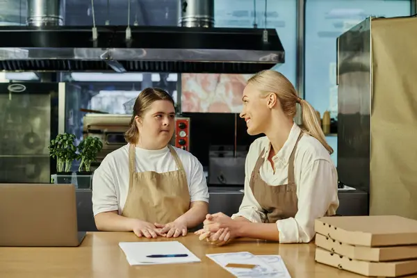 Sonriente gerente de café hablando con una joven empleada con síndrome de Down cerca de la computadora portátil y cajas de pizza - foto de stock
