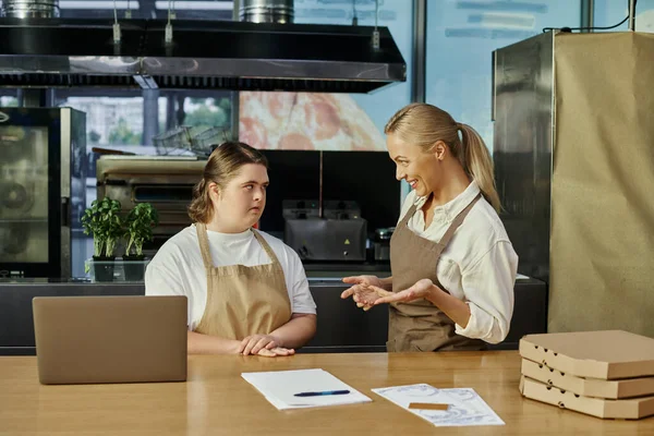 Joyous administrator explaining duties to young woman with down syndrome near laptop and pizza boxes — Stock Photo