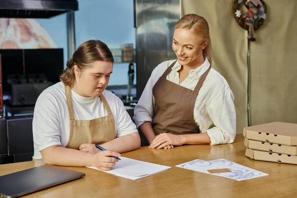 Administrador do sexo feminino sorrindo perto empregado mulher com síndrome de down escrever ordem no balcão no café — Fotografia de Stock