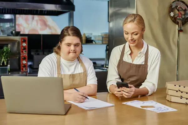 Jovem com deficiência mental escrevendo ordem perto de laptop e gerente com smartphone no café — Fotografia de Stock