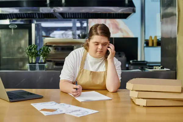 Junge Café-Mitarbeiterin mit Down-Syndrom spricht auf Smartphone neben Laptop und Pizzakartons auf Theke — Stockfoto
