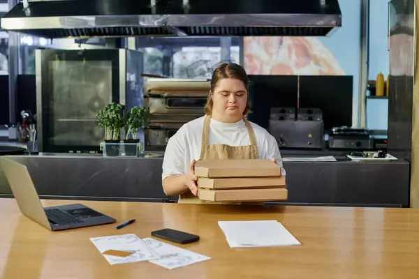 Female employee with down syndrome holding pizza boxes near laptop and smartphone on counter in cafe — Stock Photo