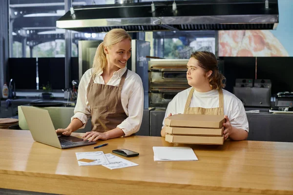 Sonriente gerente de café trabajando en el ordenador portátil cerca de empleada femenina con síndrome de Down sosteniendo cajas de pizza - foto de stock