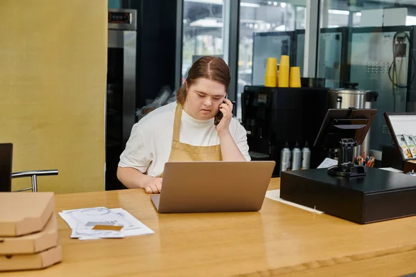 Mujer joven con síndrome de Down hablando en el teléfono inteligente cerca de la computadora portátil mientras trabaja en la cafetería moderna - foto de stock