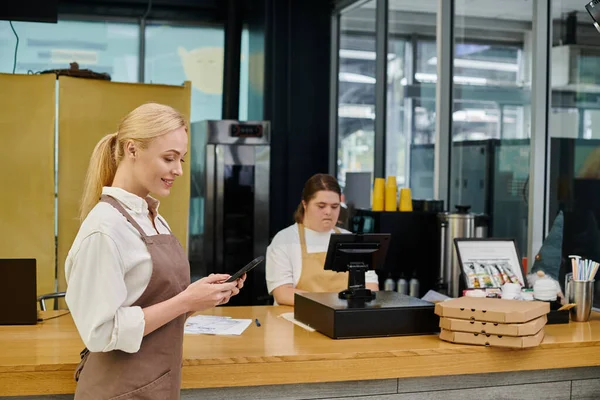 Happy cafe manager messaging on smartphone near female employee with down syndrome at cash terminal — Stock Photo