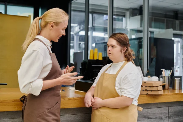 Gerente sonriente hablando con una joven empleada con síndrome de Down en el mostrador en la cafetería moderna - foto de stock