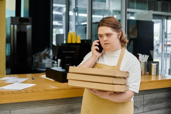 Young female employee with down syndrome holding pizza boxes and talking on smartphone in cafe — Stock Photo
