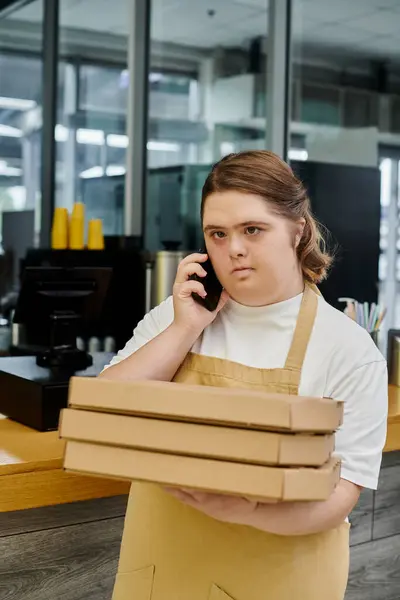 Mujer joven con síndrome de Down sosteniendo cajas de pizza y hablando en el teléfono inteligente mientras trabaja en la cafetería - foto de stock