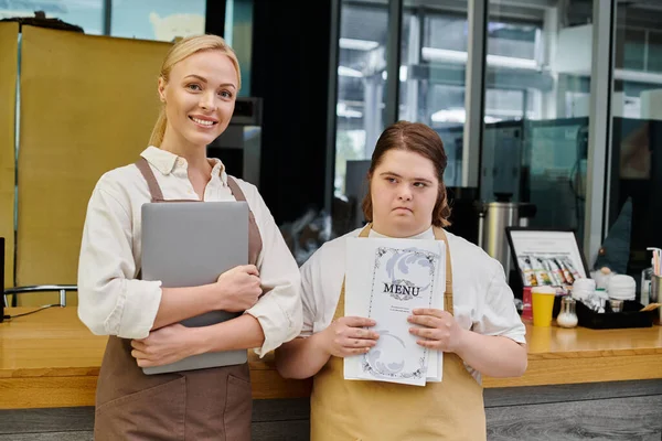 Feliz gerente y mujer joven con síndrome de Down celebración de la computadora portátil y tarjeta de menú en la cafetería moderna - foto de stock