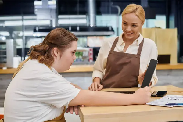 Administrador alegre mirando a la mujer joven con síndrome de Down trabajando en el ordenador portátil en la cafetería moderna - foto de stock