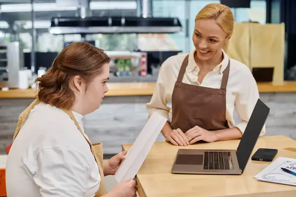 Happy manager looking at young woman with down syndrome holding order book near laptop in cafe — Stock Photo