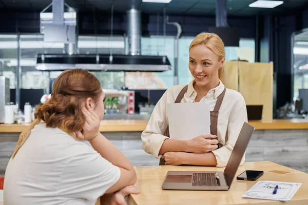 Felice gestore di caffè con libro degli ordini parlando con dipendente donna con sindrome di Down vicino laptop — Foto stock