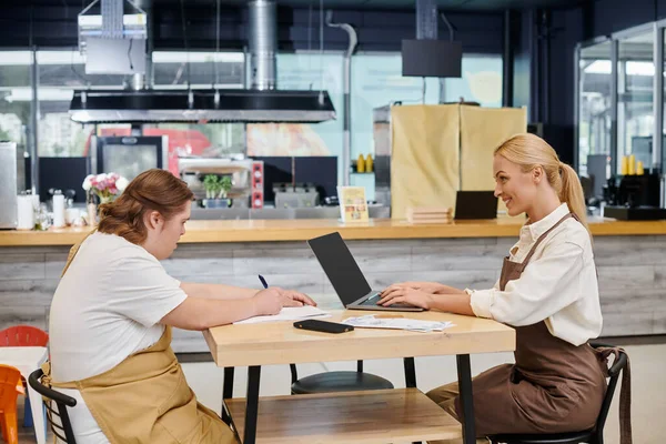 Administrador alegre que trabaja en el ordenador portátil cerca de empleada con discapacidad mental en la mesa en la cafetería - foto de stock