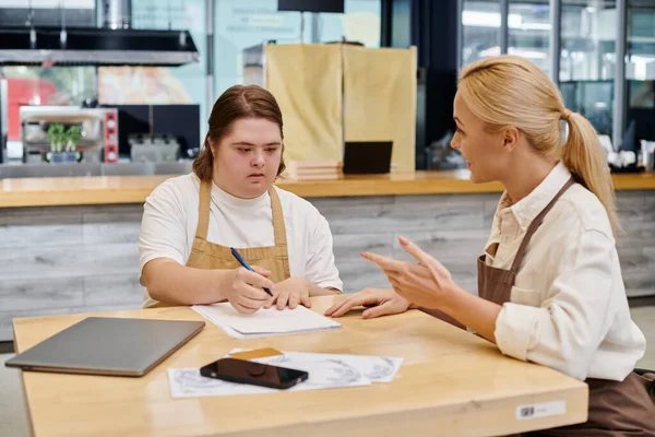 Administrador de la cafetería hablando con una empleada con síndrome de Down escribir cerca de la computadora portátil y teléfono inteligente - foto de stock