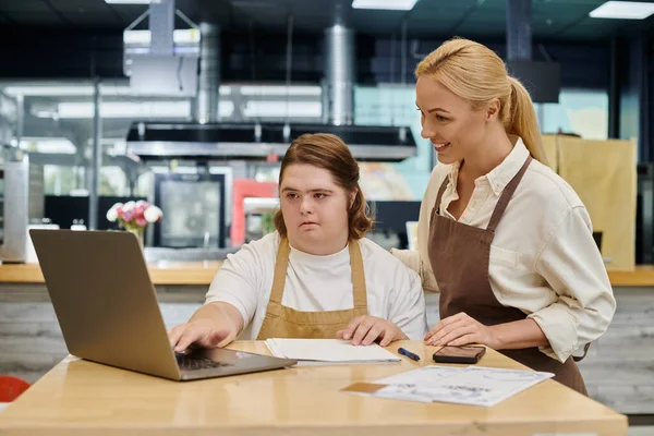Alegre administrador sonriendo cerca de empleada con síndrome de Down trabajando en el ordenador portátil en la cafetería - foto de stock