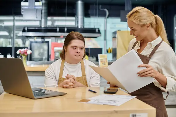 Administrador alegre sentado con libro de pedidos cerca de la mujer con síndrome de Down en el ordenador portátil en la cafetería - foto de stock