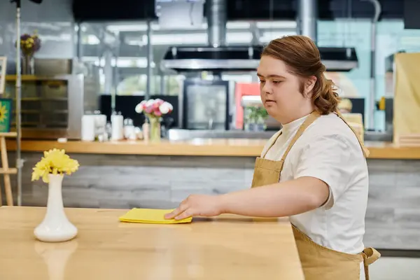 Mujer joven con síndrome de Down limpiando mesa con trapo mientras trabaja en la cafetería moderna, inclusividad - foto de stock