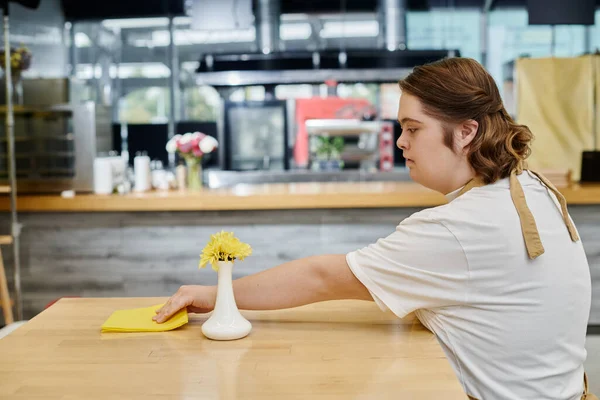 Mujer joven con trastorno mental limpiando mesa con trapo mientras trabaja en la cafetería moderna, inclusividad - foto de stock