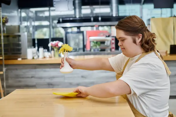 Young woman with down syndrome holding vase with flowers and wiping table with rag in modern cafe — Stock Photo