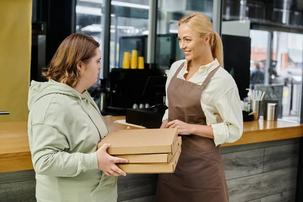 Молодая сотрудница с синдромом Дауна держит коробки с пиццей рядом с улыбающимся администратором в кафе — стоковое фото
