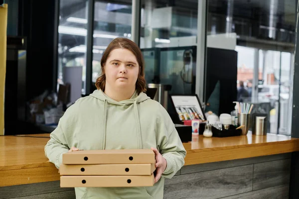 Mujer joven con trastorno mental sosteniendo cajas de pizza y mirando a la cámara en la cafetería acogedora moderna - foto de stock