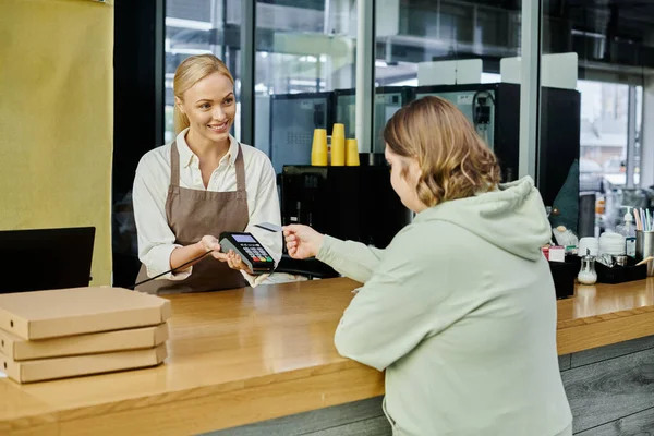 Mujer joven con síndrome de Down pagar con tarjeta de crédito cerca de administrador con terminal en la cafetería - foto de stock
