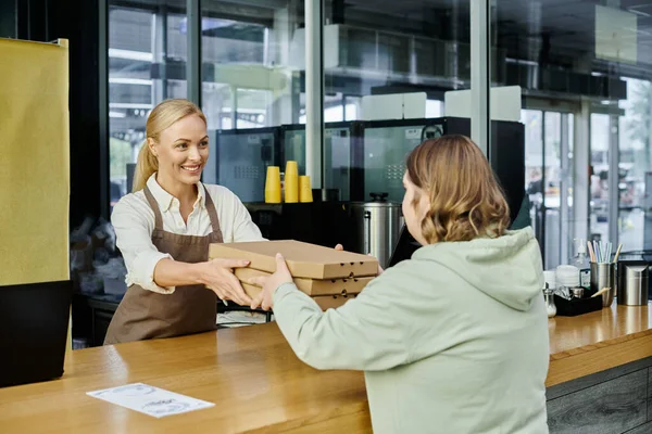 Улыбающийся администратор кафе дарит коробки пиццы клиентке с синдромом Дауна в современном кафе — стоковое фото