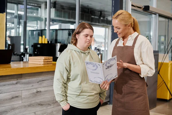 Smiling female administrator showing menu card to thoughtful woman with down syndrome in modern cafe — Stock Photo