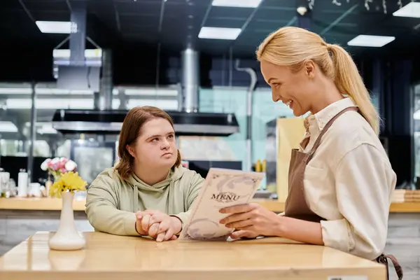 Gerente feliz mostrando la tarjeta de menú a la mujer reflexiva con síndrome de Down sentado en la mesa en la cafetería - foto de stock