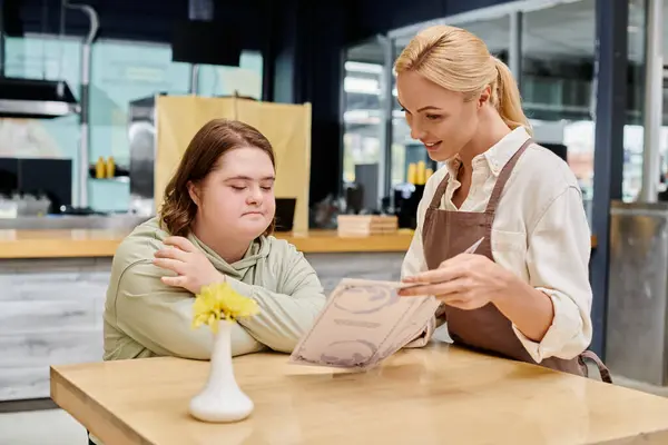 Smiling waitress showing menu card to thoughtful woman with down syndrome sitting at table in cafe — Stock Photo