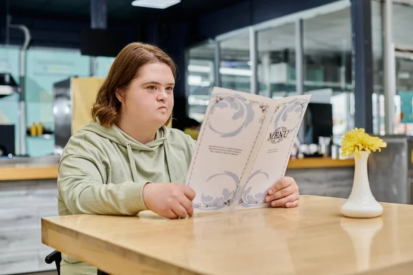 Nachdenkliche junge Frau mit Down-Syndrom schaut auf Speisekarte, während sie am Tisch im Café sitzt — Stockfoto