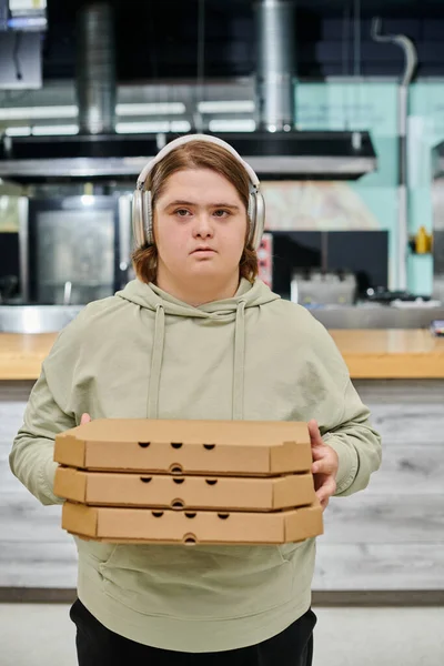 Jeune femme avec des troubles mentaux dans des écouteurs sans fil tenant des boîtes à pizza dans un café moderne — Photo de stock