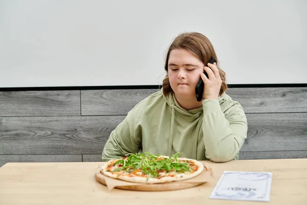 Junge Frau mit geistiger Störung spricht in modernem Café neben leckerer Pizza auf Smartphone — Stockfoto