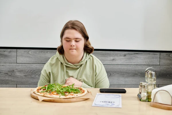 Young woman with down syndrome looking at delicious pizza while sitting in modern cozy cafe — Stock Photo