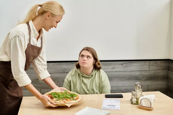 Joyful waitress proposing delicious pizza to female client with down syndrome sitting in cozy cafe — Stock Photo