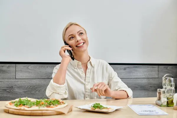 Счастливая блондинка с сообщениями на смартфоне рядом с вкусной пиццей на столе в современном уютном кафе — стоковое фото