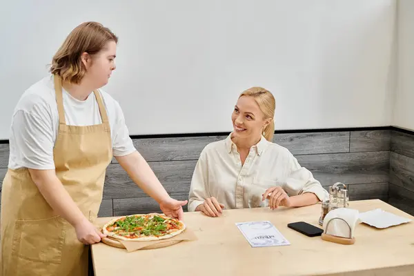 Jeune serveuse atteinte de troubles mentaux proposant une délicieuse pizza à une femme souriante dans un café moderne — Photo de stock