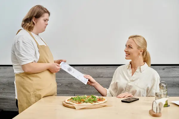 Young waitress with mental disability giving menu card to happy woman sitting in modern cafe — Stock Photo