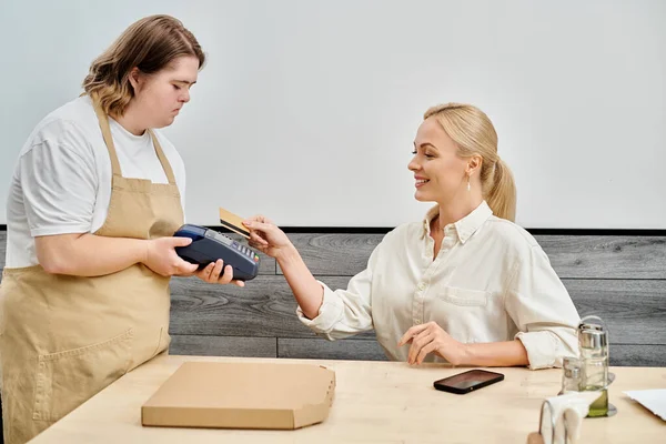 Young waitress with down syndrome holding payment terminal near pleased woman in modern cafe — Stock Photo