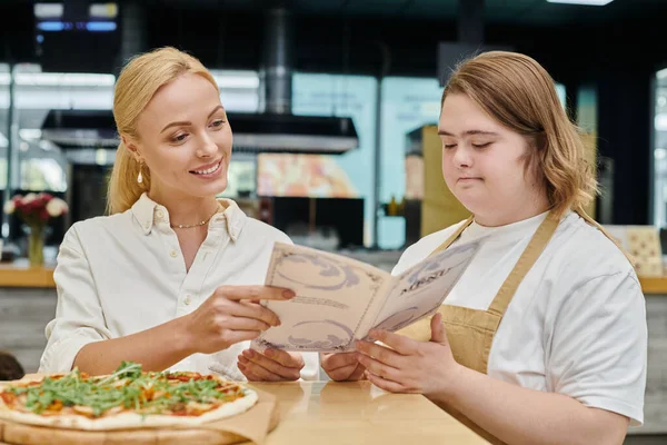 Young waitress with down syndrome showing menu card to smiling woman near pizza in modern cafe — Stock Photo