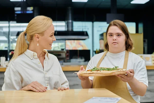 Молодая официантка с умственной отсталостью держит вкусную пиццу рядом с радостной женщиной, сидящей в кафе — стоковое фото
